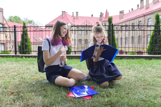 Two schoolgirls sitting on grass with books near school building, girls sisters, teenager and elementary school student laugh and look at textbooks