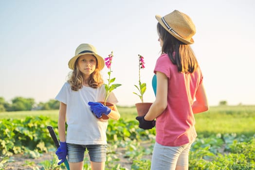 Portrait of two girls children with flowers in pots, gloves, with garden shovels. Young gardeners in hats, background rural nature, landscape, sky, spring summer season