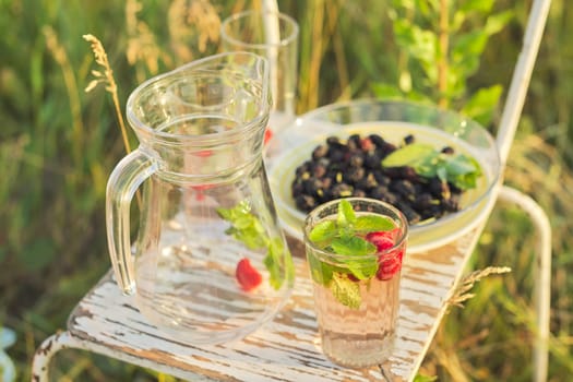 Summer natural vitamin berry drinks, jug and glasses with drink strawberry mint, plate with mulberry, background of summer herbs flowers on lawn, sunny day