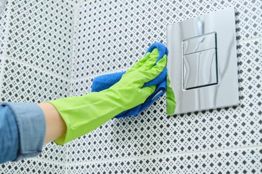 Woman in gloves with rag doing cleaning in bathroom, cleaning and polishing chrome toilet button on tiled wall