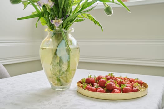 Home dining room interior, table with white tablecloth, spring summer bouquet of flowers in vase, tray with ripe strawberries
