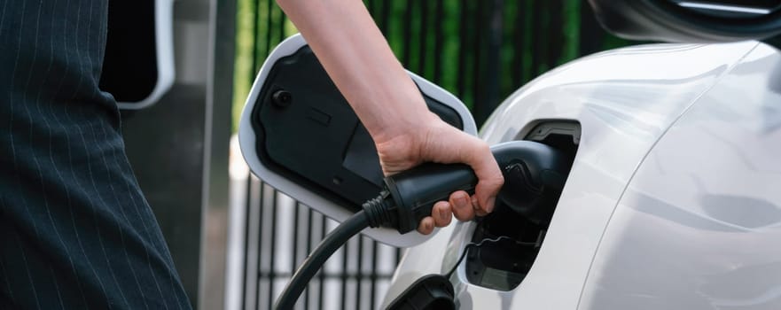 Focus EV charger plug and electric car at public charging station with blur progressive businesswoman holding charger and apartment condo building in background. Eco friendly electric vehicle concept.