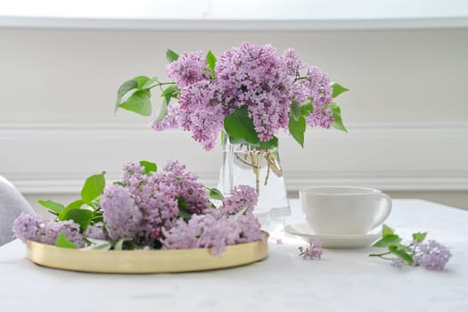 Spring bouquet of lilac flowers in glass jug on table, cup of tea, spring holidays, sunny mood, background white tablecloth