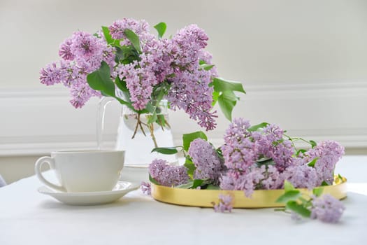 Spring bouquet of lilac flowers in glass jug on table, cup of tea, spring holidays, sunny mood, background white tablecloth