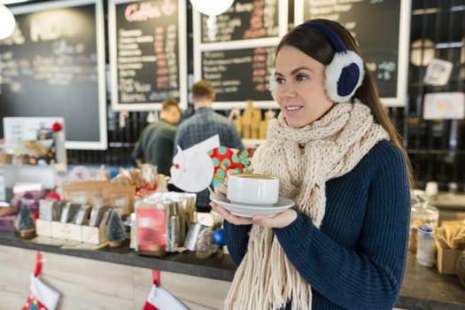 Winter portrait of young beautiful woman in knitted scarf, fur ears, warm sweater with cup of coffee. Girl standing near bar counter in coffee shop