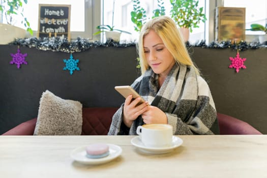 Beautiful blonde girl in woolen blanket with smartphone in the interior of winter coffee house, macaroon in plate, cup of coffee