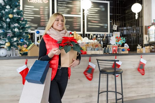 Happy smiling blonde in coffee shop with shopping bags and red poinsettia christmas flower. Winter portrait, girl in woolen scarf, with Christmas shopping