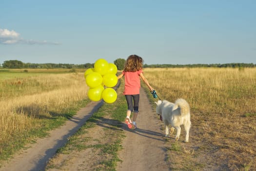 Dynamic outdoor portrait of running girl with white dog and yellow balloons on country road, beautiful landscape with blue sky and yellow grass in meadow