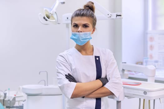 Portrait of confident female dentist doctor with folded arms in white coat with mask near dental chair in dental clinic office
