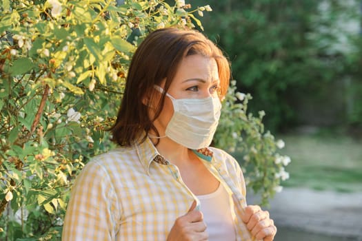 Mature woman in protective medical mask, female outdoor near flowering bush, pollen allergy