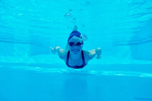 Young girl in swimsuit with goggles and swimming cap swimming underwater in the pool, female looking and smiling at the camera