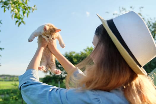 Teenager girl in country style denim hat holding little ginger kitten in her arms, nature sky background