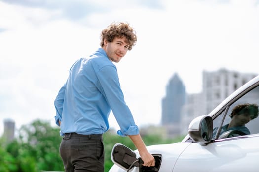 Progressive businessman insert charger plug from charging station to his electric vehicle with apartment condo building in background. Eco friendly rechargeable car powered by sustainable energy.