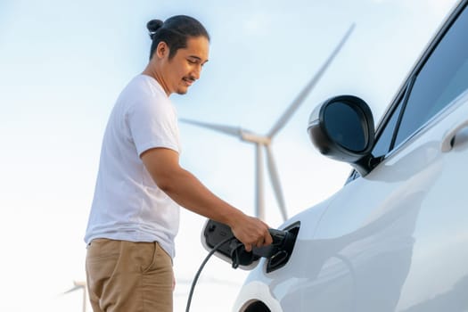 Progressive man with his electric car, EV car recharging energy from charging station on green field with wind turbine as concept of future sustainable energy. Electric vehicle with energy generator.