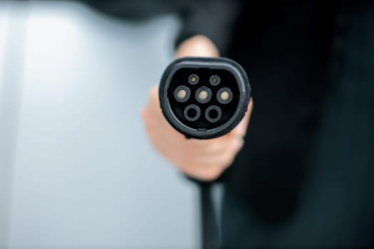 Focus closeup hand holding EV plug for electric vehicle pointing at camera with charging station at background as progressive idea of alternative sustainable clean energy for ecological awareness idea