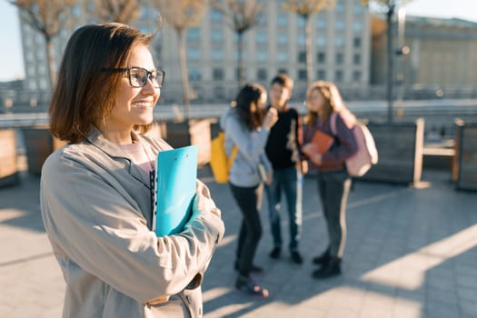 Portrait of mature smiling female teacher in glasses with clipboard, outdoor with a group of teenagers students, golden hour.