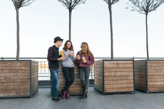 Teenagers eat street food, friends boy and two girls on city street with burgers and orange juice. City background, golden hour