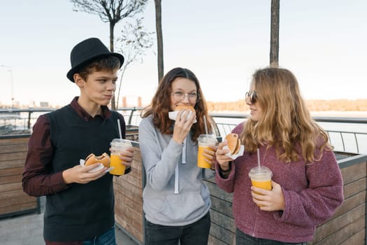 Teenagers eat street food, friends boy and two girls on city street with burgers and orange juice. City background, golden hour