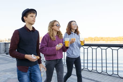 Lifestyle of adolescents, boy and two teen girls are walking in the city. Laughing, talking children eating street food, having fun. Background of the river embankment