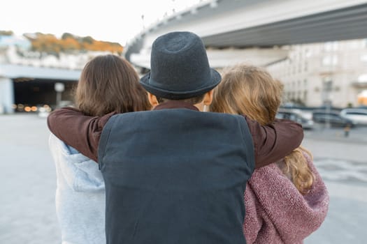 Three friends, view from the back of young teenager boy hugging two girls by the shoulders. Children look ahead, city life