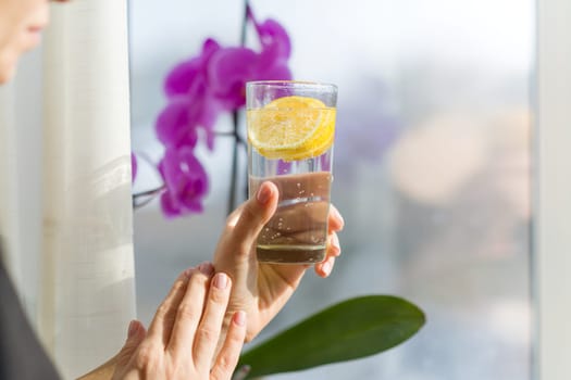 Mature woman is holding a glass with healthy drink. Natural antioxidant water with lemon, female stands near a window, enjoys drink.