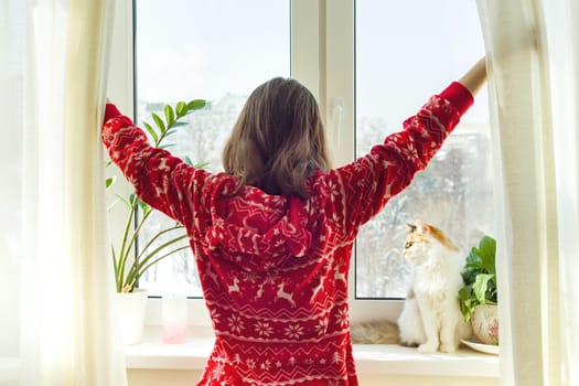 Winter time at home, young girl in winter warm pajamas with a cat looking out the window.