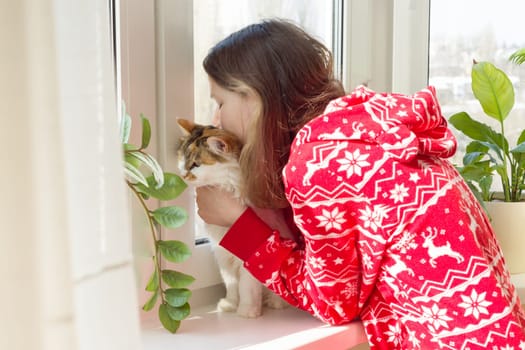 Winter time at home, young girl in winter warm pajamas with a cat looking out the window.