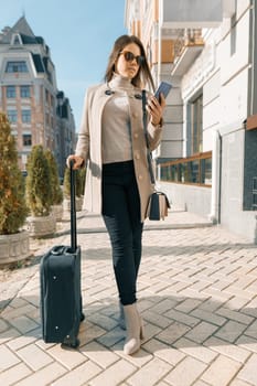 Portrait of traveling young woman with mobile phone and suitcase, fashionable girl on the city street, wearing warm coat, sunny autumn day.