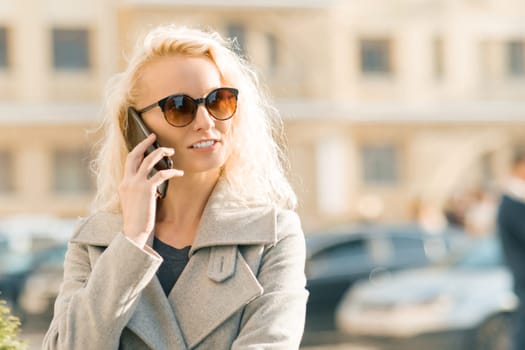 Outdoor portrait of young blonde woman with curly hair smiling and talking on a mobile phone on sunny autumn day in the city.
