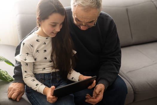 Happy family senior grandfather and girl watching cartoon on tablet on weekend day at home together.