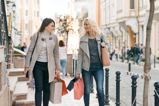 Urban style, two young smiling fashionable women walking along a city street with shopping bags, sunny autumn day, golden hour.