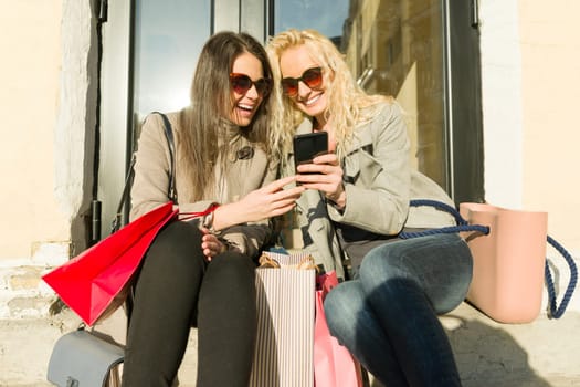 Two young smiling women on a city street with shopping bags, sunny autumn day, golden hour.