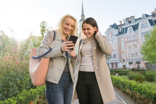 Two young women having fun, looking at the smartphone laughing, sunny autumn day, city background, golden hour.