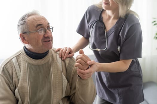 Caring nurse talks to old patient holds his hand sit in living room at homecare visit provide psychological support listen complains showing empathy encouraging. Geriatrics medicine caregiving concept.