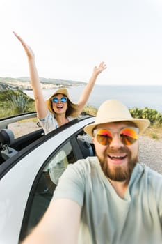 Happy beautiful couple in love taking a selfie portrait driving a convertible car on the road at vacation. Rental cars and vacation concept