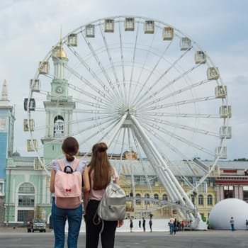 Two young girls with backpacks looking at the Ferris wheel, back view, in the city.