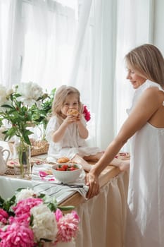 A little blonde girl with her mom on a kitchen countertop decorated with peonies. The concept of the relationship between mother and daughter. Spring atmosphere