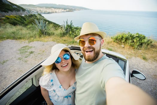 Romantic couple making selfie on smartphone camera in rental cabrio car on ocean or sea beach, enjoying summer vacation together and taking picture cellular resting near sea on weekends
