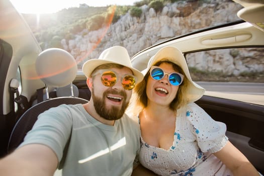 Happy beautiful couple in love taking a selfie portrait driving a convertible car on the road at vacation. Rental cars and vacation concept