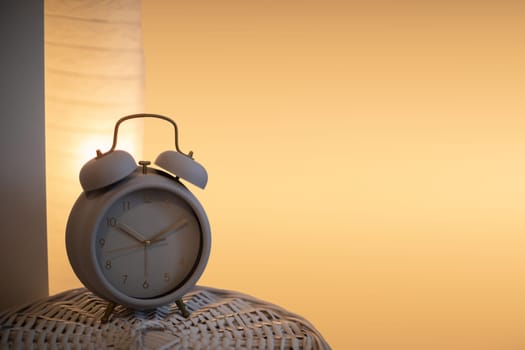 A lit bedside lamp gives a soft orange-yellow light to the background and an analog alarm clock stands in the foreground. The evening atmosphere of the bedroom.