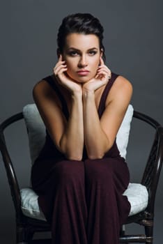 Fashionable beautiful young woman with hairstyle and makeup posing on a dark background. Female in trendy knitted overalls on a wicker chair