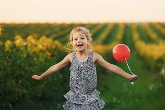 Positive little girl with red balloon in hands have fun on the field at summer day time.