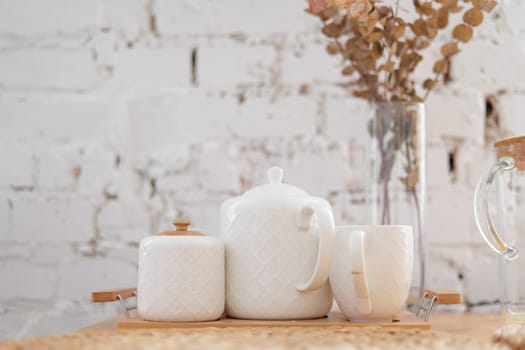 The tea white set on wooden tray on a white brick wall background. Teapot, creamer, cup and glass vase with dry eucalipt branches on the table. Ceramic kitchenware. English afternoon tea set. Studio.