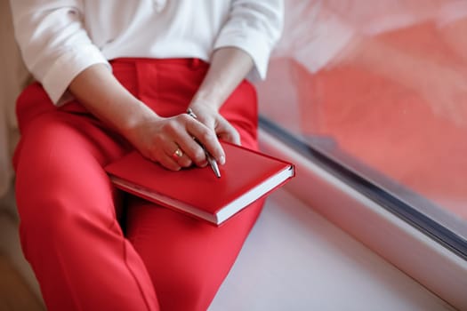 cropped photo of young European businesswoman with notebook is sitting by the window. Concept of planning. girl in white shirt and red pants