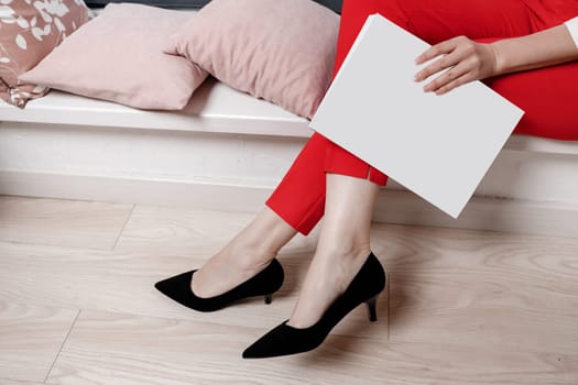 cropped photo of woman hands holding a magazine with mock up. girl in red pants and high heeled shoes sitting on sofa.