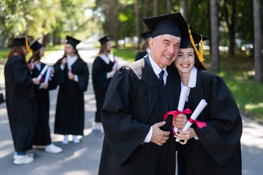 A group of graduates in robes outdoors. An elderly man and a young woman congratulate each other on their graduation