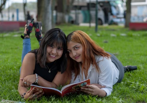 two friends lying on the grass in a park smiling and reading a book. High quality photo