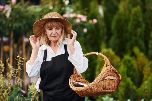 With basket in hands. Senior woman is in the garden at daytime. Conception of plants and seasons.