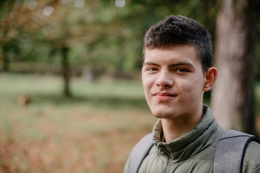 a guy in a jacket stands on an alley in the park during the fall season. Portrait oh handsome teen guy, young man in hoodie, down jacket standing, walking in beautiful golden autumn park, looking at camera. Natural background, colourful leaves, trees. Portrait of young smiling man in casual jacket looks at camera on autumn nature background in countryside or in park. Concept of style, walking in fresh air and unity with nature