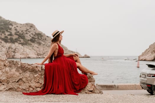 Side view a Young beautiful sensual woman in a red long dress posing on a volcanic rock high above the sea during sunset. Girl on the nature on overcast sky background. Fashion photo